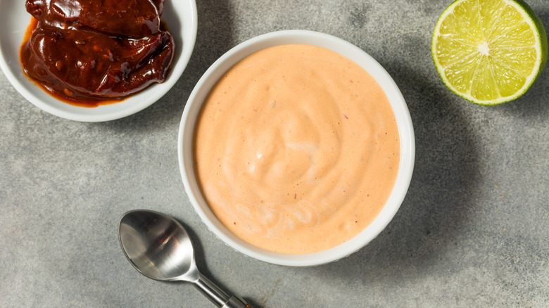 bowl of homemade chipotle mayo dip next to a bowl of chipotle peppers and a halved lime