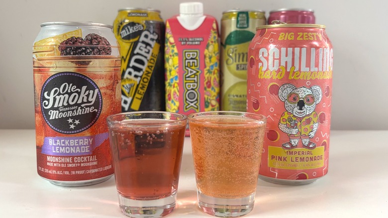 Two shot glasses of hard lemonade between two cans of hard lemonade with others in the background, on white table