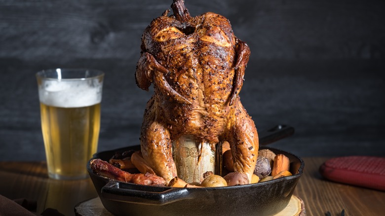 A cooked beer can chicken sitting on the can in a pan with different vegetables with a glass of beer in the background