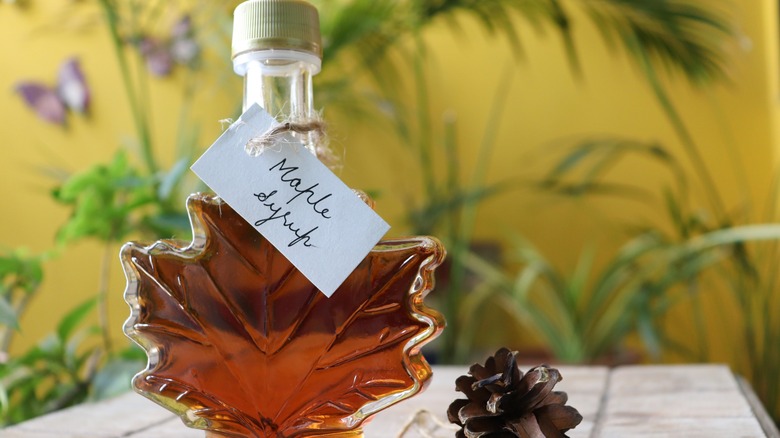 Maple leaf shaped bottle filled with maple syrup standing on a white table with a pine cone on the side and greenery in the back