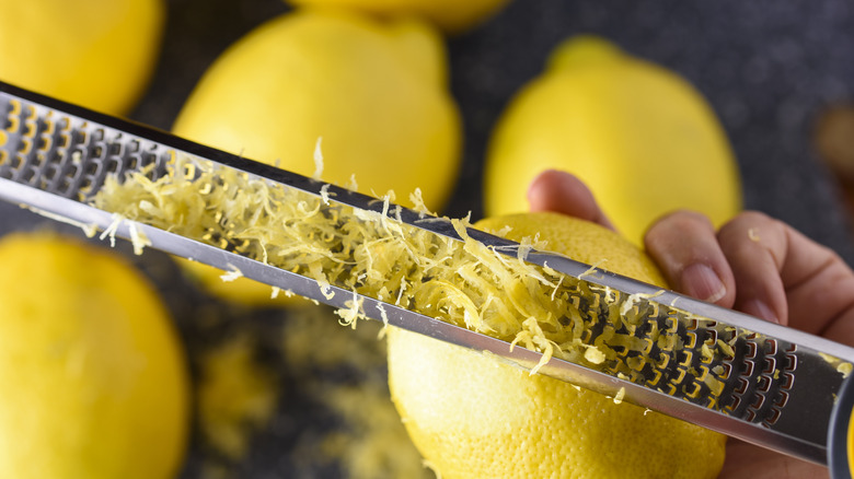 A person zesting a lemon with a metal grater with lemons in the back