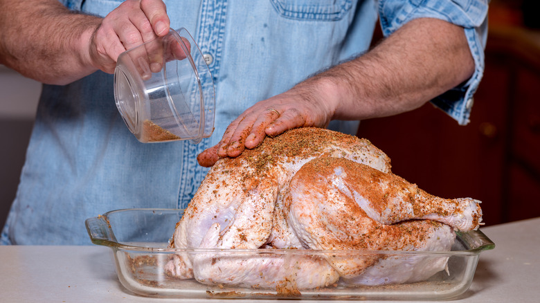 A person patting dry spices onto a raw whole turkey that is nestled in a glass pan