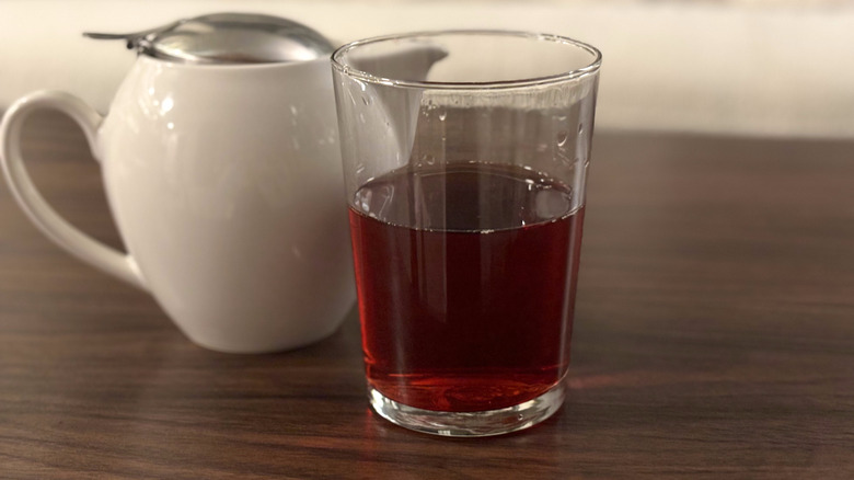 Clear glass with dark red hibiscus tea inside in front of small white teapot on table