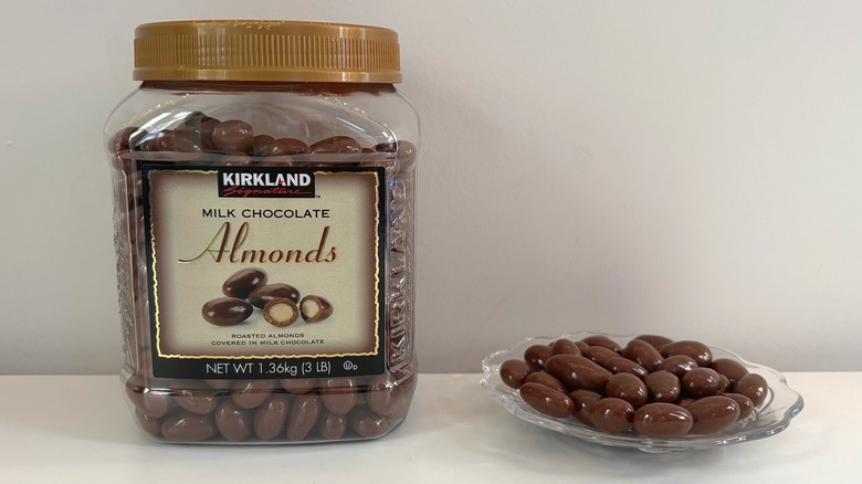 Container of Kirkland milk chocolate almonds next to glass plate of chocolate almonds on white background