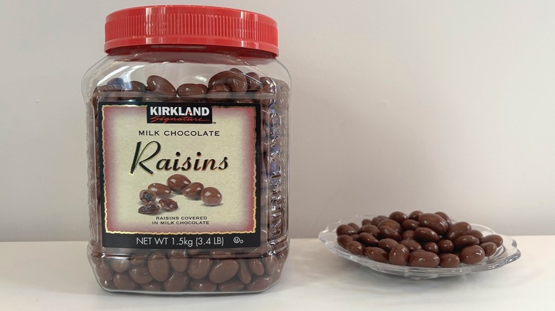 Container of Kirkland milk chocolate raisins next to glass plate of milk chocolate raisins on white background