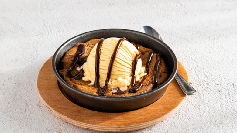 Chili's Skillet Chocolate Chip Cookie topped with a scoop of vanilla ice cream and a drizzle of chocolate sauce, all in a grayish-black bowl on a round wood plate with a metal spoon