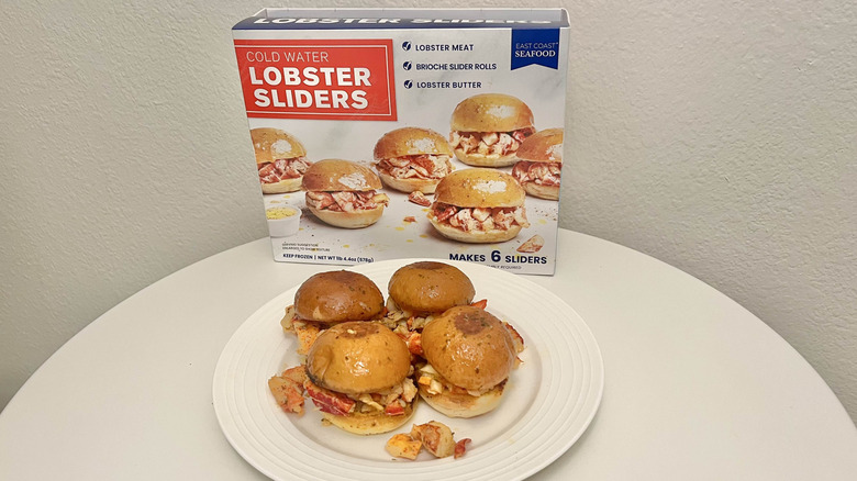 Four East Coast Seafood lobster sliders sit on a plate with the product's packaging behind it.