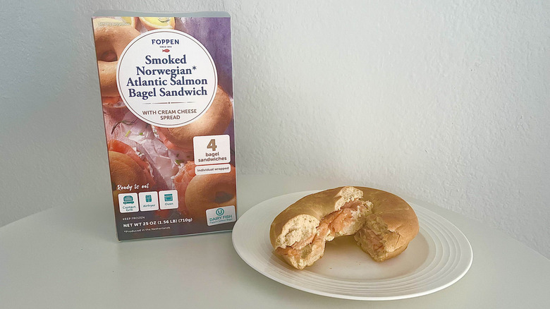 A Foppen smoked salmon bagel sandwich is cut in half on a plate with the product's packaging behind it.