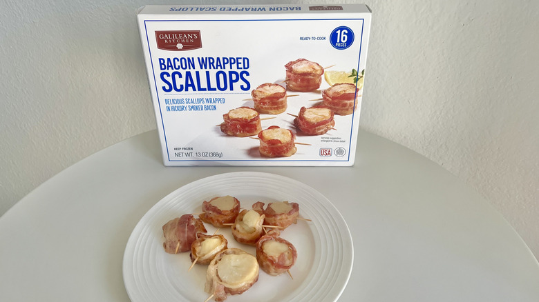 Bacon-wrapped scallops are arranged on a white plate with the product's packaging behind it.