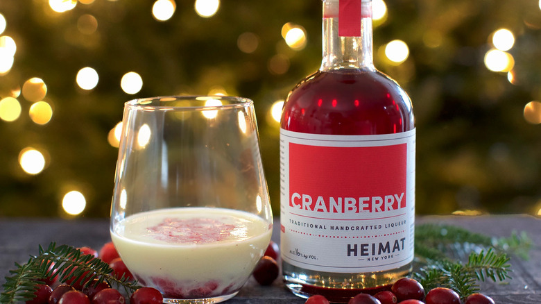 Bottle of Heimat liqueur next to a festive egg nog cocktail, surrounded by festive lights and fresh cranberries.