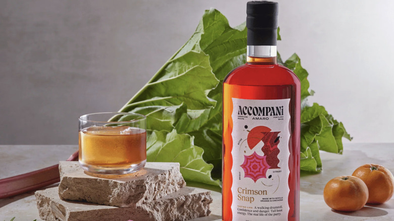 A bottle of Accompani's bright red Crimson Snap liqueur, posed with a mixed drink, a pair of oranges, and a stalk of rhubarb.