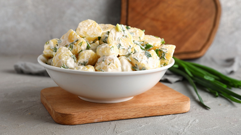 Bowl of potato salad balanced on wooden cutting board with chives in the background.