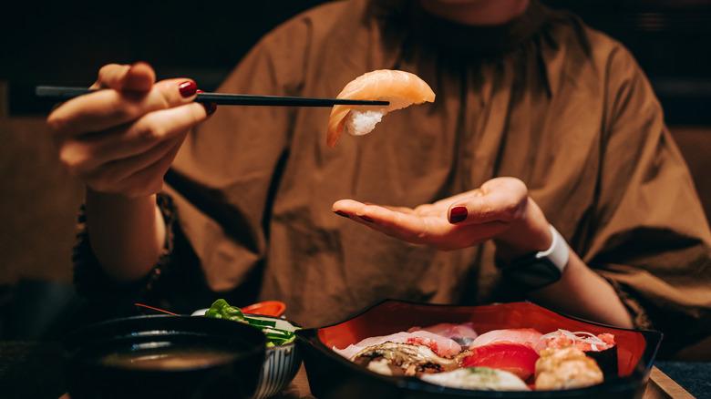 A woman holding sushi with chopsticks, a bowl of assorted sashimi and a bowl of soup in front of her