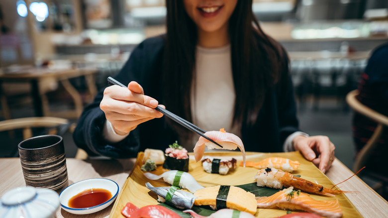 Woman smiling while holding up sashimi over a tray of assorted sashimi with chopsticks