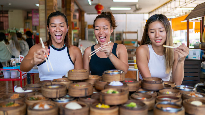 Three women holding chopsticks in front of a pile of bamboo baskets with dim sum