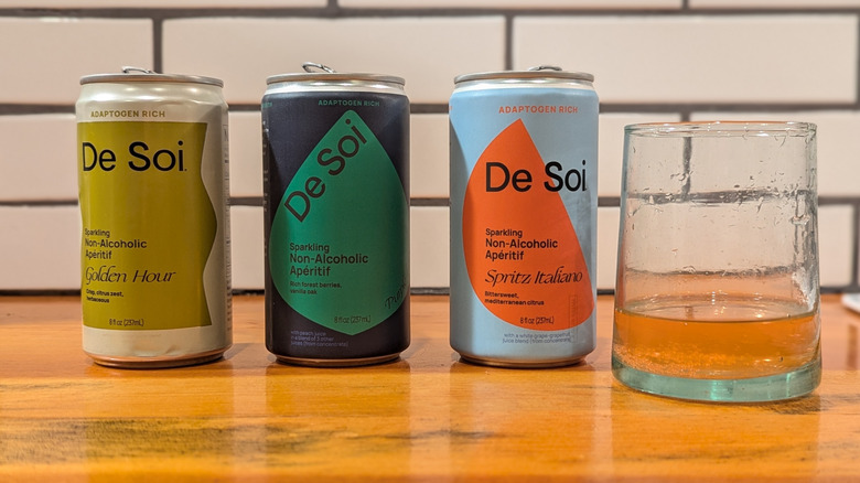 three cans of aperitif drinks with colorful, geometric labels and a glass with drink