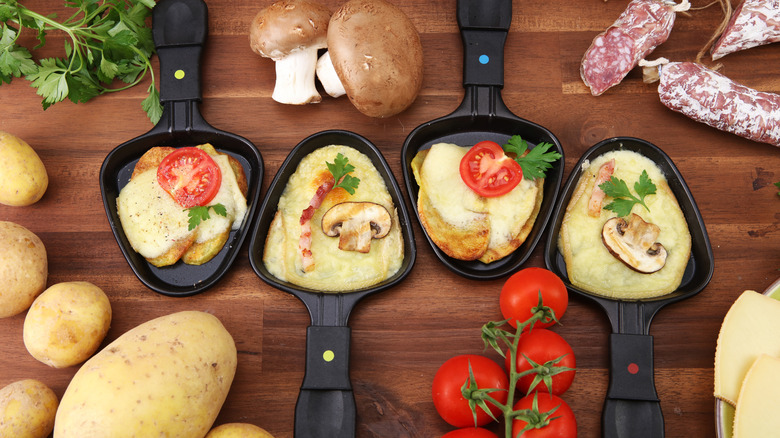 four raclette spatulas lined up on a wooden counter filled with cheese and different toppings and surrounded with fresh parsley, potatoes, mushrooms, tomatoes, sausage, and cheese