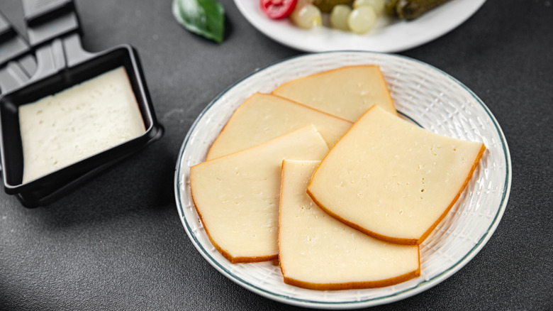 slices of raclette cheese assembled on a white plate with a raclette grill tray and plate of veggies in the back