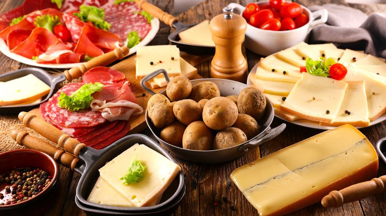 a table spread with plates and spatulas packed with raclette cheese next to a block of cheese, bowl full of potatoes, peppercorns, charcuterie, and cherry tomatoes