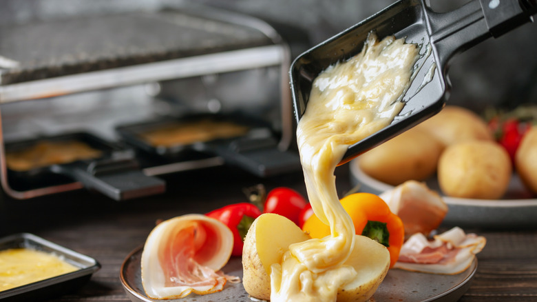 pouring melted raclette over boiled potatoes placed next to prosciutto slices and veggies with a raclette grill in the background