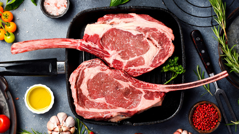 two raw tomahawk steaks on frying pan surrounded by herbs and seasonings