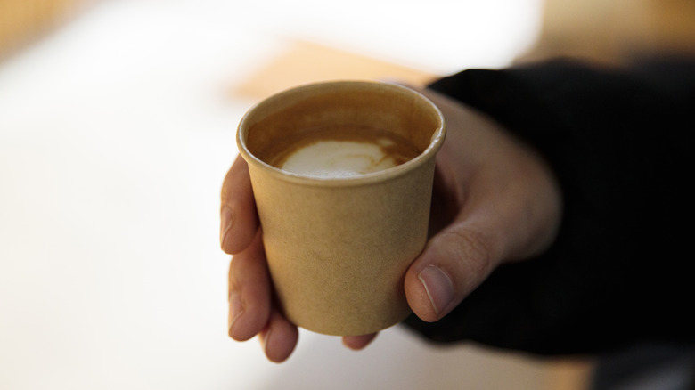 Person holding a small cup of frothy coffee in a brown paper cup