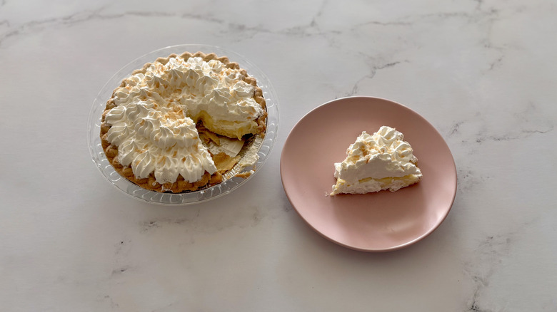 A coconut cream pie sitting next to a slice of coconut cream pie that is on a plate.