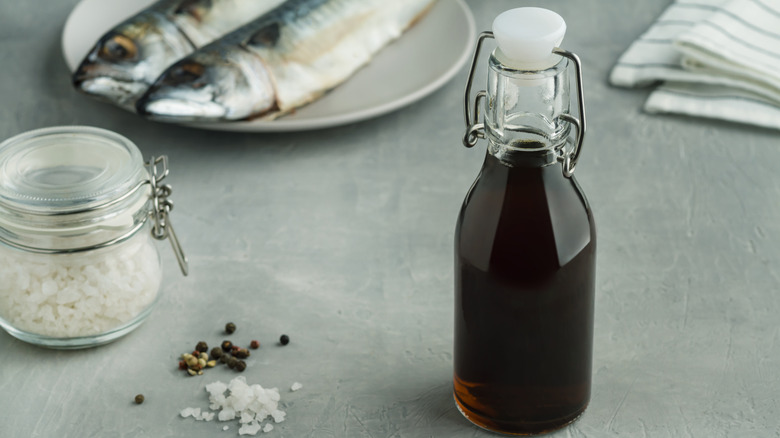A glass bottle of fish sauce next to a glass jar of salt with a plate of fish in the background