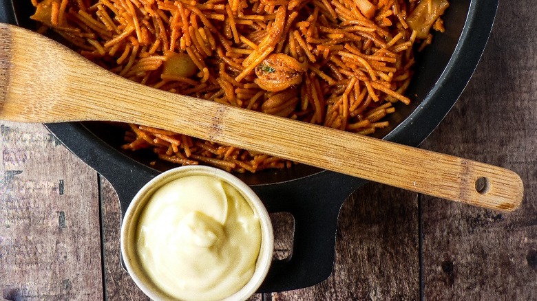 Fideua in a cast iron pan with a wooden spoon and a ramekin of aioli on a wooden tabletop