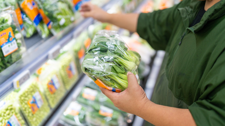 Person holding bagged produce wrapped in plastic, with more plastic bagged produce on shelves