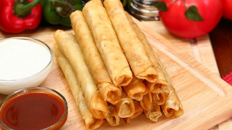 A stack of flour tortilla wrapped flautas with tomatoes, hot sauce, and Mexican crema