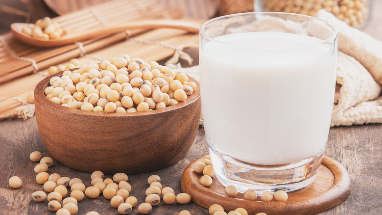 a glass of soy milk next to a bowl of soy beans on a table with a bamboo place mat, napkin, and wooden spoon