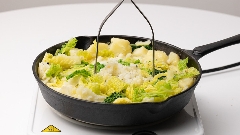 potato masher over cabbage and potatoes in cast iron pan