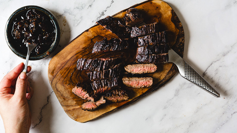 Hand spooning sauce out of bowl besides cutting board with steak