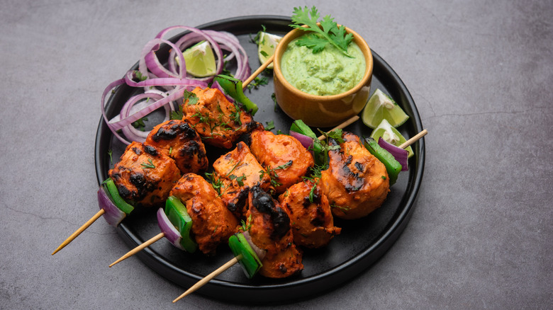 chicken tikka skewers on a plate with a bowl of sauce and some garnishes of onion and lime