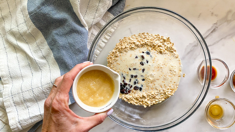 Hand adding applesauce to bowl of oats, milk, and raisins