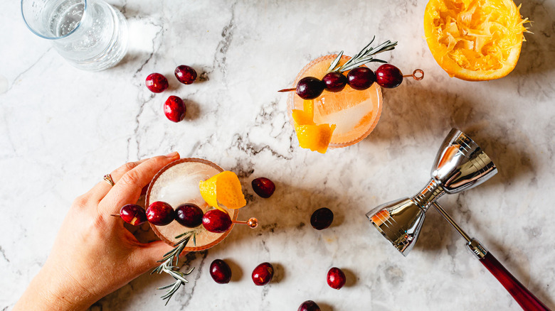 hand holding one of two cocktail glasses with jigger, cranberries and orange half