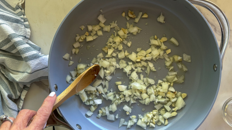 Hand stirring onions and garlic in a pan with a wooden spoon