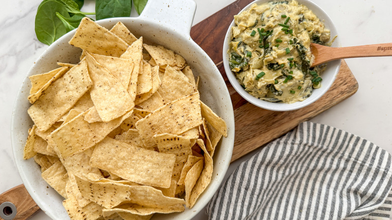 Chip and spinach artichoke dip in white bowls on cheese board