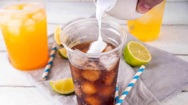 Plastic cup of soda with cream being poured in surrounded by beverages, lime, and straws