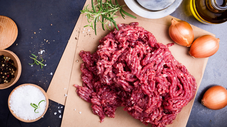 Ground beef surrounded by shallots, salt and pepper, rosemary and olive oil with a mortar and pestle in the background