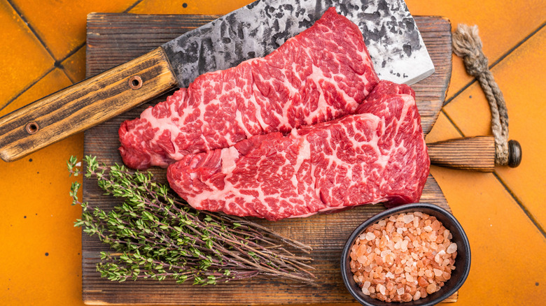 Raw Denver steaks on cutting board with herbs, salt, and a cleaver.