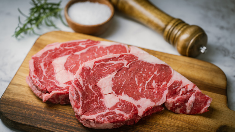 Raw ribeye steaks on wooden cutting board near salt and herbs.