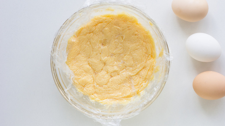 Pastry cream in a glass bowl covered in plastic wrap on a counter with three eggs on the side sitting on a white counter