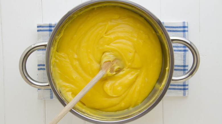 Thick and creamy yellow pastry cream in a pot with a wooden spoon on a white surface covered in a kitchen towel