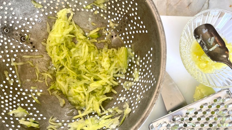 A metal colander with shredded cucumber next to a glass bowl and a shredder  