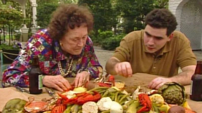 Emeril Lagasse and Julia Child hunched over vegetables on table