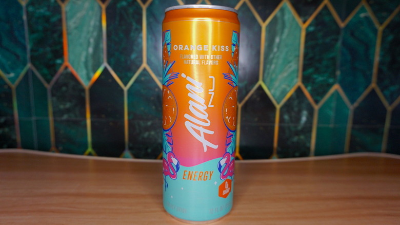 Orange Kiss Alani Nu energy drink on a wooden table, against a green-and-gold tiled background