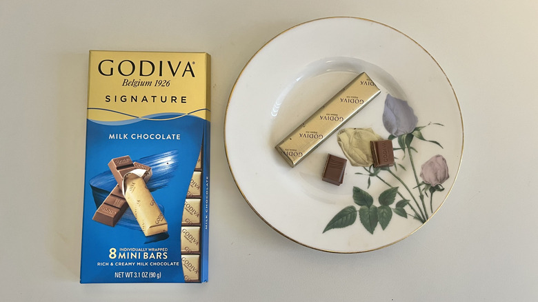 A Godiva milk chocolate mini bar sits on a plate next to the package on a table.