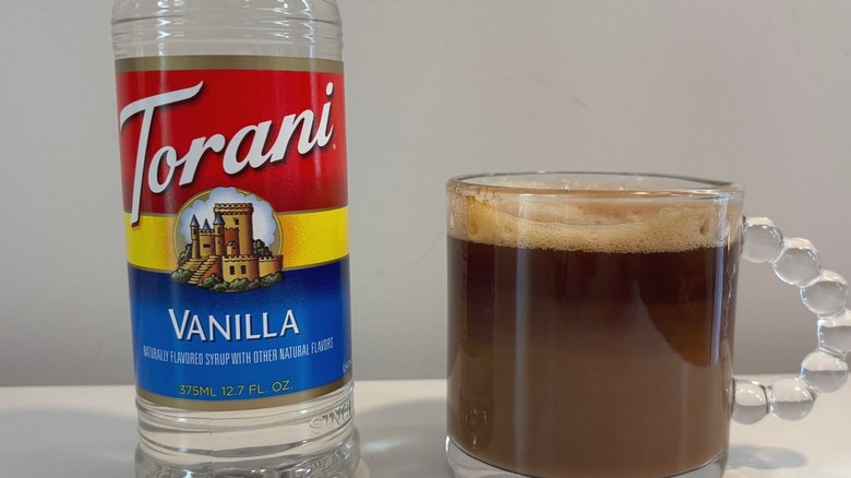 Clear mug of Nespresso coffee with oat milk next to bottle of Torani vanilla syrup